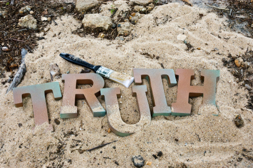 The word TRUTH spelled out in metal letters on a sandy beach next to a paintbrush