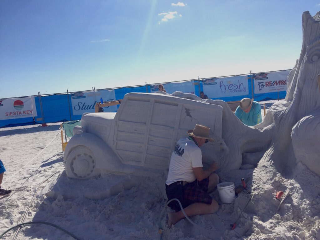 Sand sculptor carving a detailed truck at a beach sand-sculpting competition