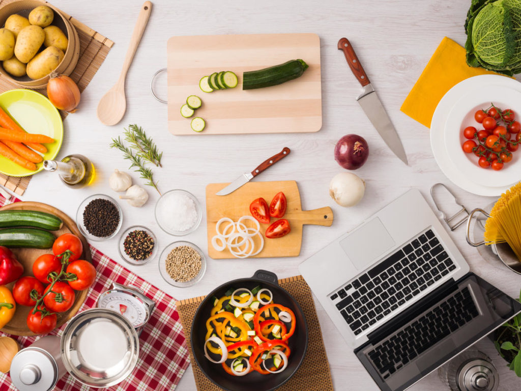 Overhead view of chopped vegetables, cooking ingredients, and a laptop arranged on a kitchen counter
